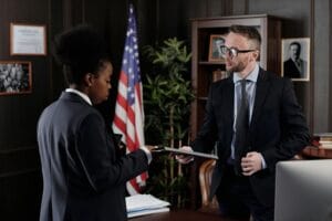 Business professionals engage in a meeting in a formal office environment with an American flag in the background.