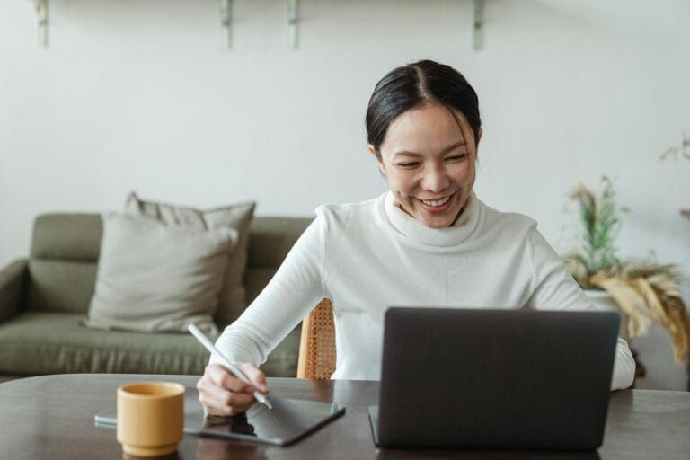 How To Find Virtual Remote Jobs Without Falling For Scams 1 A cheerful woman uses a laptop and tablet for a video call, working remotely in a cozy living room.