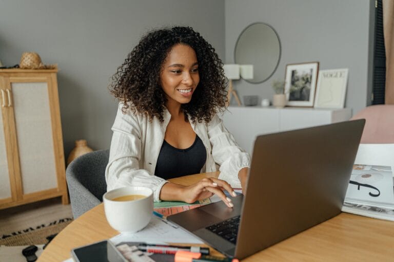 Best Staffing Agencies For Legit Remote Jobs 2 A woman sits at a round table, working on a laptop with a coffee cup nearby in a cozy home office setting.