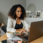 A woman sits at a round table, working on a laptop with a coffee cup nearby in a cozy home office setting.