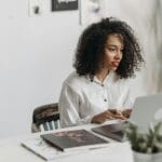 An African American woman with curly hair works attentively on a laptop in a modern office setting.