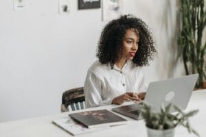 An African American woman with curly hair works attentively on a laptop in a modern office setting.