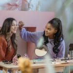 Positive African American female beauty influencer recording makeup tutorial on cellphone while applying eyeshadow on client