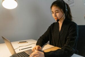 Asian woman using a laptop with a headset in a modern office setting.