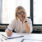 Smiling woman talking on the phone while writing notes at a desk in a modern office.