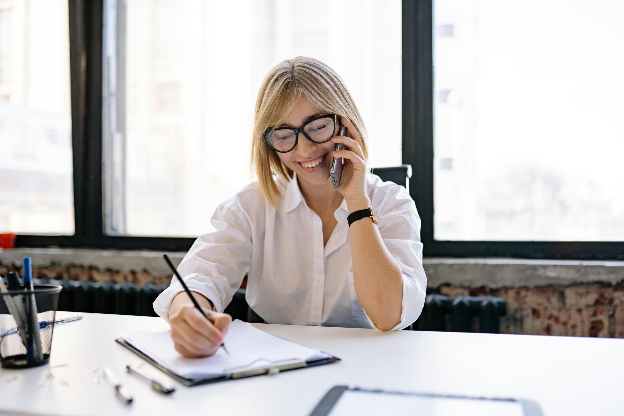 Smiling woman talking on the phone while writing notes at a desk in a modern office.