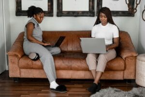 Two African American women sitting on a couch, working from home on laptops.