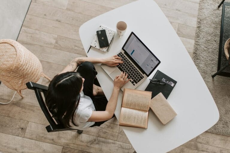 Overhead View Of A Woman Using A Laptop At A Home Desk, Surrounded By Books, A Phone, And A Cup.