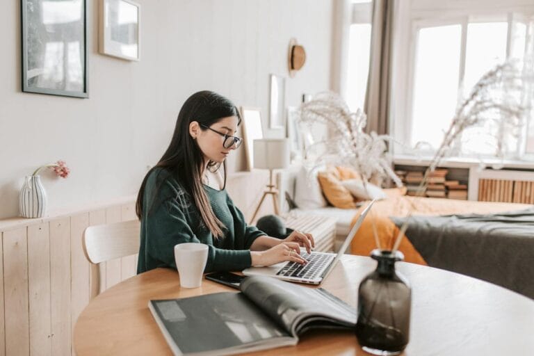 Side View Of Female Remote Worker In Eyeglasses And Casual Wear Typing On Netbook While Sitting At Wooden Table With Cup And Open Magazine In Bedroom At Home