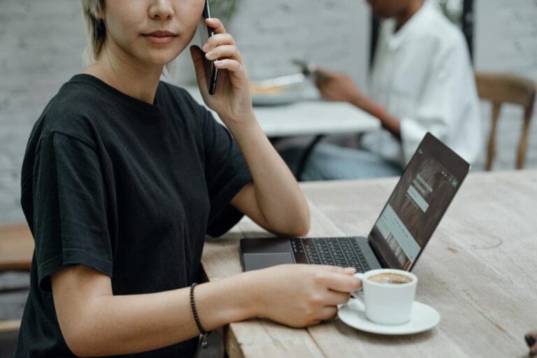 Crop Asian Lady In Black Tee Making Phone Call While Sitting At Table With Coffee And Laptop In Cafe