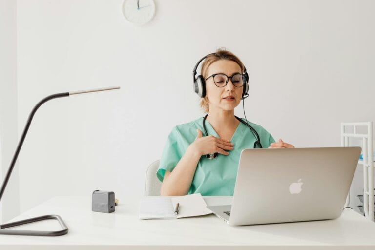 Doctor In A Green Scrub Conducting An Online Video Consultation Using A Laptop And Headphones.