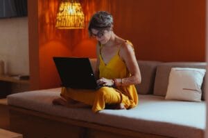 Senior Woman In Comfortable Attire Using A Laptop In A Warm, Indoor Setting, Showcasing Modern Technology And Leisure.