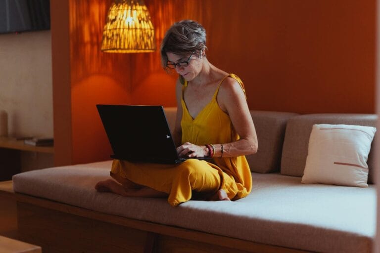 Senior Woman In Comfortable Attire Using A Laptop In A Warm, Indoor Setting, Showcasing Modern Technology And Leisure.