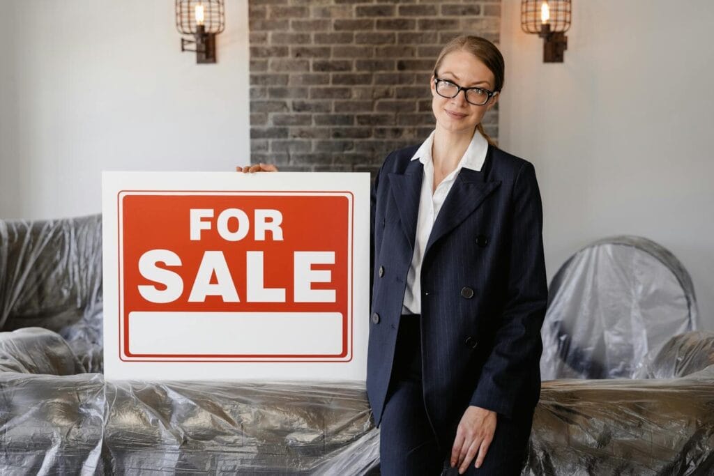 Real Estate Agent In Formal Attire Holding A Blank 'For Sale' Sign Inside An Apartment.