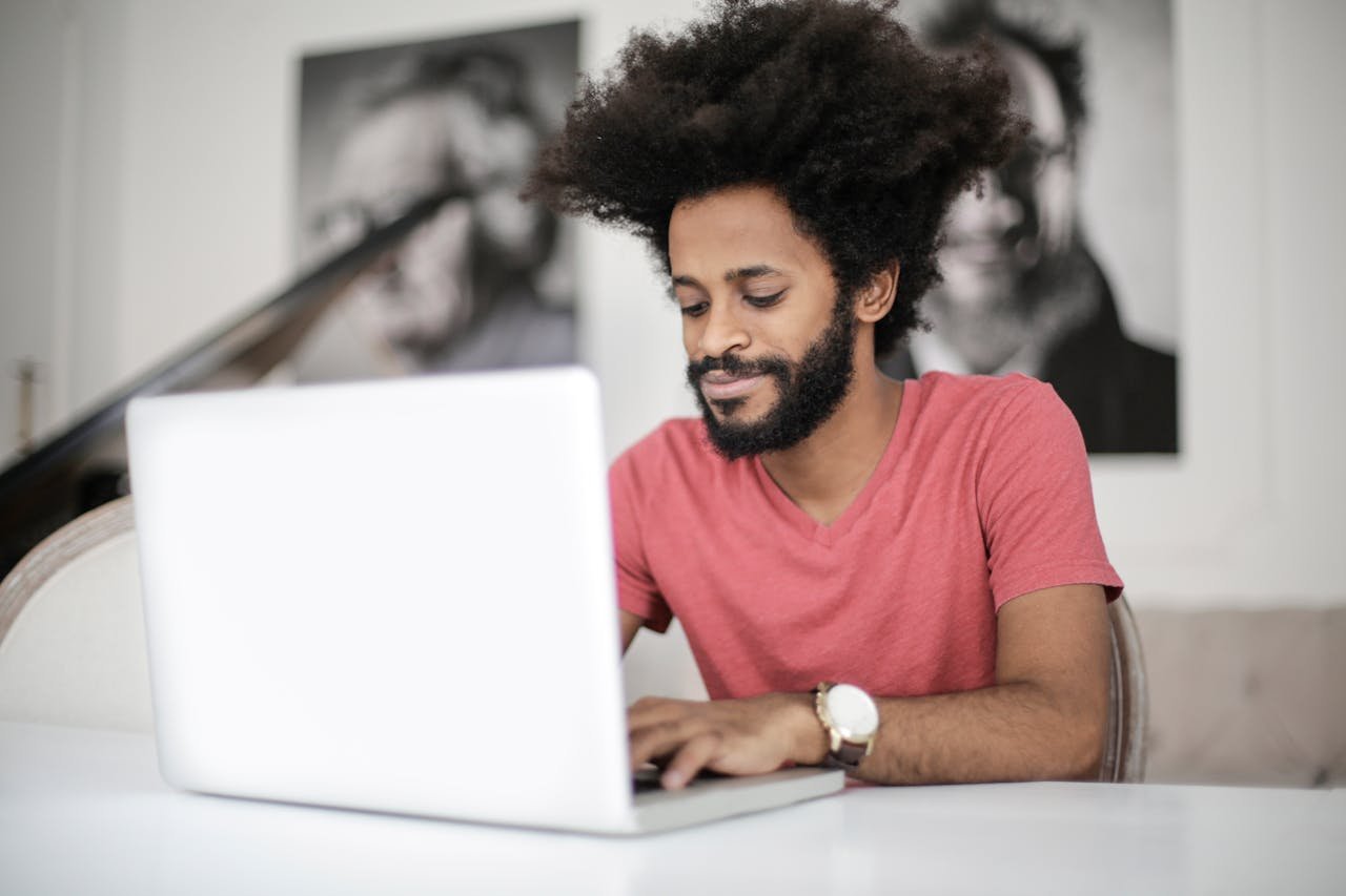 A Man With Afro Hair Works On His Laptop At A Desk, Symbolizing Modern Remote Work And Freelance Lifestyle.