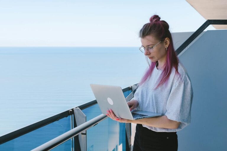 Is Becoming A Virtual Assistant Worth It? (2026 Complete Guide) 9 A Young Woman Works Remotely On Her Laptop By The Sea On A Sunny Day, Embodying The Digital Nomad Lifestyle.