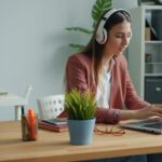 Focused young woman wearing headphones while working remotely on her laptop from a home office.
