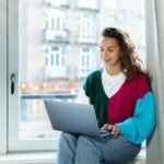 a woman sitting on a window sill using a laptop