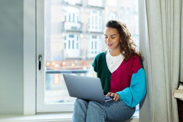 A Woman Sitting On A Window Sill Using A Laptop